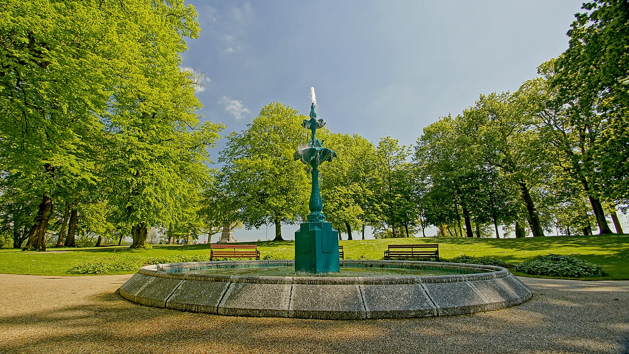 Green fountain surrounded by paths, benches and trees in Lisburn Castle Gardens